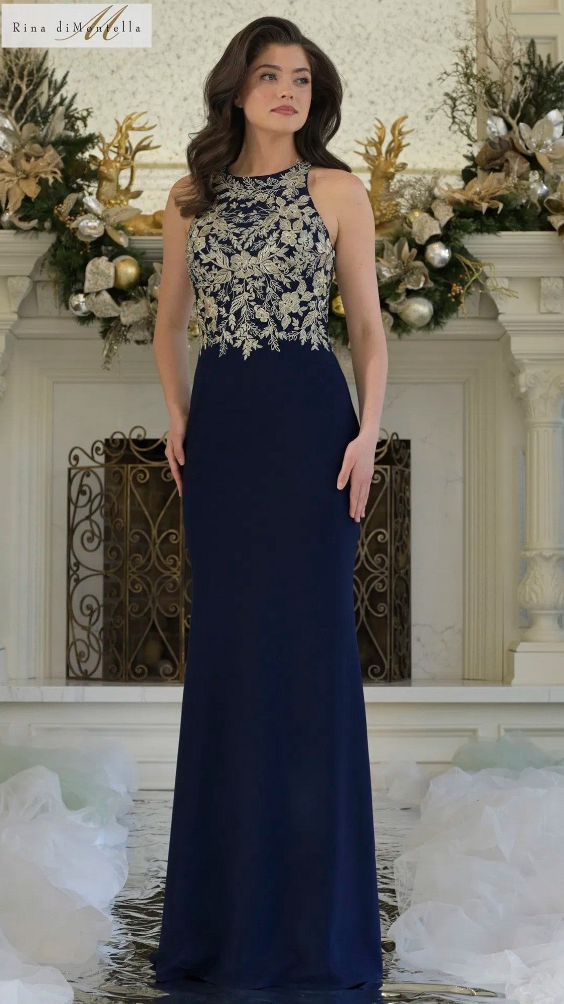 A woman stands indoors in the Rina Di Montella RD3345 long formal dress, a sleeveless black gold gown with intricate embroidery on the bodice, in front of a decorated white fireplace.